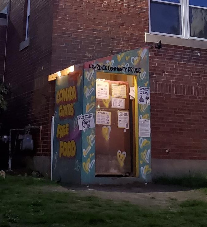 a painted shed with a diaganol sloped roof situated on the lawn in front of a brick wall. inside the shed is a refrigerator, with various signs posted on the fridge and the shed. above the fridge on the shed are hand painted words 'Lawrence Community Fridge' and the mural on the side of the shed reads in big yellow letters: 'free food, comida gratis.