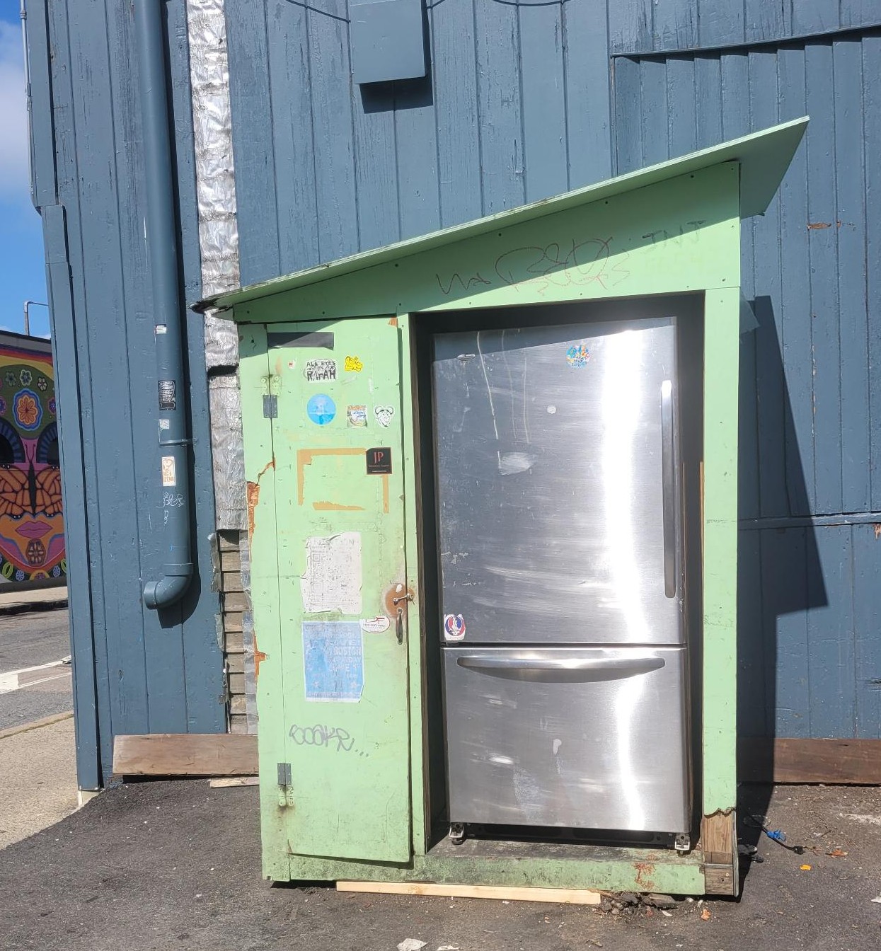 a shed painted bright green housing a stainless steel fridge. both are outside on the pavement against a blue paneled exterior wall. there is a closed cabinet door to the left of the fridge.