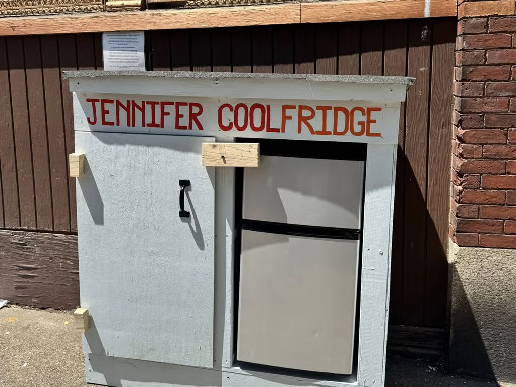 a small refrigerater inside of a white shed against a brick wall. next to the fridge is a closed cabinet door with a black handle. red text above the fridge reads 'JENNIFER COOLFRIDGE'